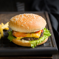 Delicious fresh tasty burger with beef, tomato, cheese, lettuce and french fries served on a wooden cutting board on dark background. Street fast food