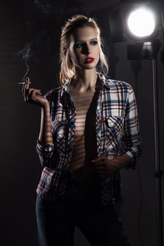 Beautiful smoking slender blonde big breasts girl without a bra wearing an unbuttoned checkered shirt and jeans covered with shadows from the blinds near the studio lamp