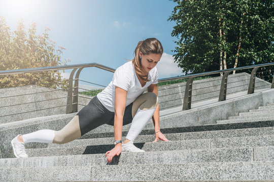 Sunny summer day. Young woman in sportswear doing stretching exercises outdoor. Girl doing warm-up on steps before training. Exercise in street, sports exercises, workout. On background green trees.