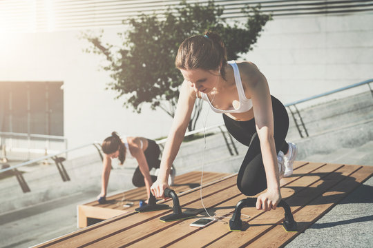 Sunny summer day. Two young smiling women, girls in sportswear doing exercises while listening to music. Workout, training, couching on city street.