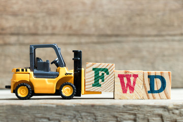 Toy yellow forklift hold letter block F to complete word FWD (Abbreviation of forward)on wood background