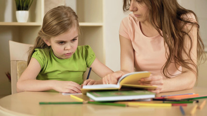 Upset little girl doing school tasks with mother, bored or tired of homework
