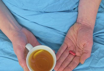 Ill man with fever drinking cup of warm tea and taking pill. Influenza season. Hands of the sick man holding pill and cup of the hot tea in the bed. Close-up of ill caucasian man at home