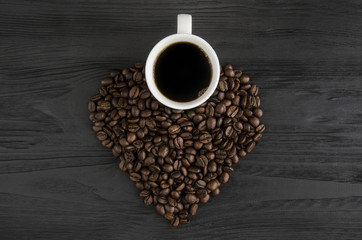 A cup of coffee and fried coffee beans in the form of a heart on a black wooden background.