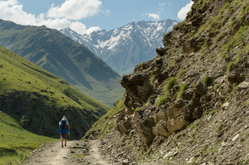 Fototapeta na wymiar Backpacker girl walking along the road. Rocky hill on the foreground, high mountain reidge partially covered with snow on the background. Hot summer day at Caucasus mountains at Tusheti region, Georgi