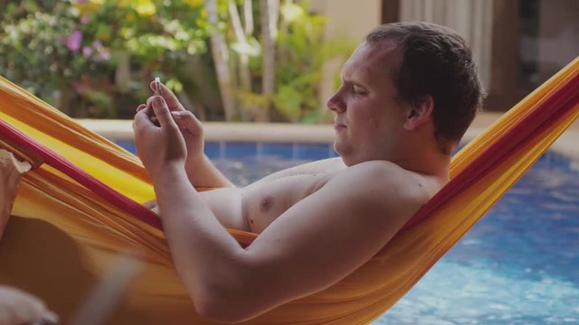 Fat lazy man with bare chest lies in hammock uses mobile phone read the book by swimming pool. 3840x2160