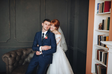 Fototapeta premium The red-haired bride in a lace dress and her groom are embracing in the interior. Cabinet with books.e translucent cloth. Wedding day.