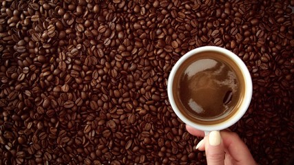 top view of hand with cup of black coffe put it on coffe beans background. White cup of black coffee