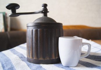 An old coffee grinder and a white cup on a white tablecloth with a geometric print