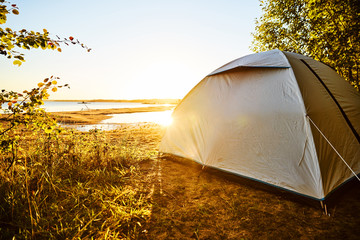 White tent standing at a beach camping spot at lake Vänern in Sweden. The sun is shining and soon will be sunset. Tent is standing on the right side covered by trees  and plants. Water reflections.  
