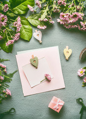 Greeting preparation with blank paper card mock up with heart and gift box on florist desktop with green leaves and flowers , top view, flat lay
