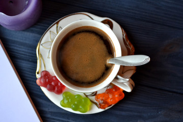 A cup of coffee with jelly candies, bright candles, pen and opened notebook on the dark blue wooden table.