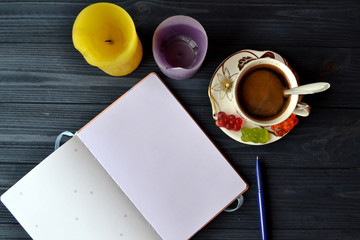 A cup of coffee with jelly candies, bright candles, pen and opened notebook on the dark blue wooden table.