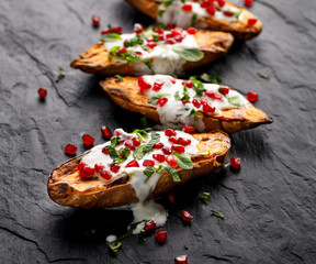 Baked sweet potatoes with garlic mint yogurt sauce sprinkled with pomegranate seeds and fresh mint leaves on a black stone background . Delicious and healthy vegetarian meal