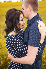 Happy young couple hugging on yellow meadow. Girl with long brunette hair in black dress in white peas, macho in blue shirt in pattern