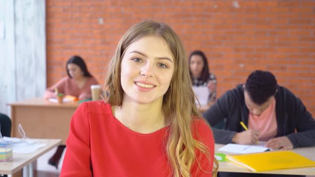 young attractive blond woman sitting opposite computer and smiling happily at camera. good-humored face of a student. In the background, young people work 4k
