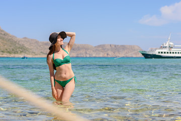 Frontal portrait of a young woman brunette in a green swimsuit standing in the emerald sea water and a hand holding a hat, copy space