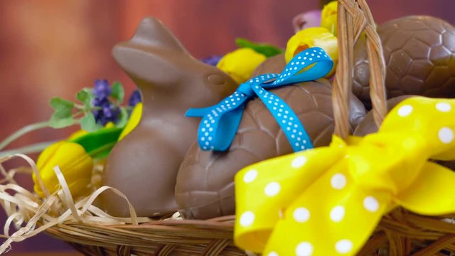Happy Easter hamper of chocolate eggs and bunny rabbits in large basket with silk tulips on dark wood table, dolly close up.