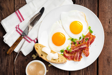 Fried eggs, bacon and italian ciabatta bread on white plate. Cup of coffee. Breakfast. Top view. Wooden background