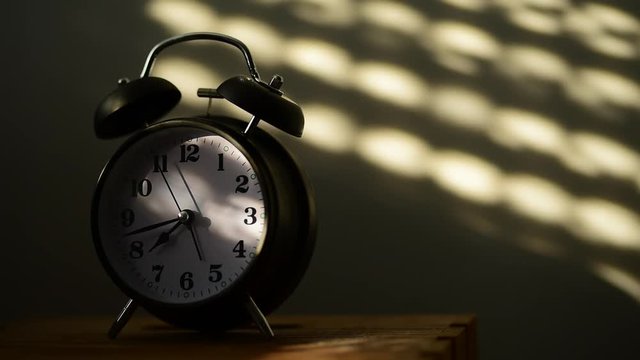 Alarm clock on night table in bedroom ticking time in early morning with sunlight and shadows on the wall in background