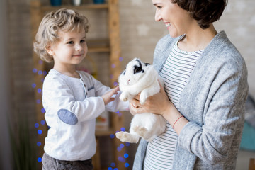 Portrait of cute curly haired blond boy playing with white pet bunny while celebrating Easter at...