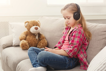 Happy little female child and her teddy bear listening to music on sofa at home