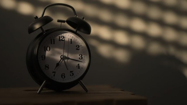 Alarm clock on night table in bedroom ticking time in early morning with sunlight and shadows on the wall in background