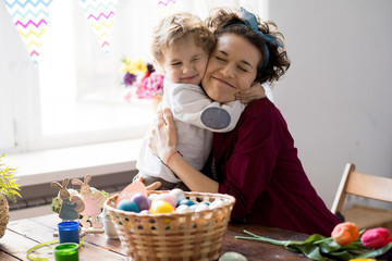 Portrait of pretty young mother hugging little boy tightly with eyes closed while celebrating Easter at home, copy space