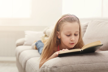 Pretty feamle kid reading book, lying on sofa at home