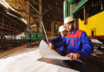 Two workers in construction helmets discuss a plan, blueprint or industrial project in the background of the plant
