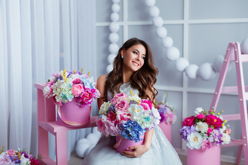 Attractive young woman in elegant dress sitting in studio interior decorated with flowers. Beautiful fresh smiling girl posing with flowers