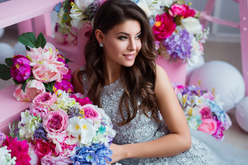Attractive young woman in elegant dress sitting in studio interior decorated with flowers. Beautiful fresh smiling girl posing with flowers