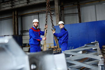 Naklejka na ściany i meble Two workers in white construction helmets load industrial products on the hook of the crane against the background of production