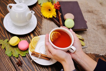 women's hands hold a cup of tea with lemon on a wooden background and against a backdrop of macaroons, old books and burlap