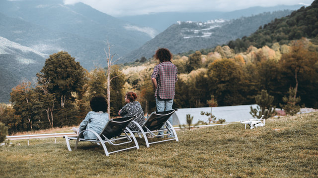 Rear view of the group of three people on the hill enjoying autumn landscape from high point of lookout: two girls are sitting on daybeds, the curly guy standing, the drone is laying on the ground