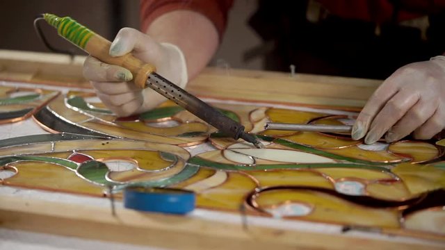 Master is using a soldering iron for decorating and processing frame of stained-glass picture. He is melting tin and applying it on copper base.