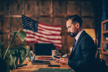 Side view image of confident enterpreneur sitting at desk and reading message on smartphone.