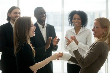 Funny smiling businesswomen holding hands celebrating success while diverse team applauding, female executive handshaking congratulating employee with common goal achievement, women power in business