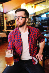 Portrait of a bearded handsome man in a red checkered shirt with glasses and a glass of beer in his hands against the background of a pub