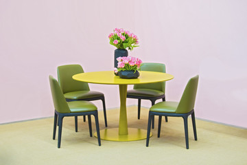 laconic interior of the dining room. Yellow round table and green leather chairs, vase with flowers on the table. Isolate on pink background