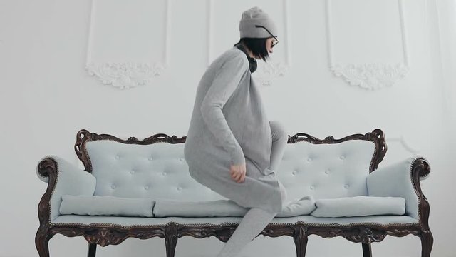 A beautiful girl or a woman dressed in gray long blouse and long gray stockings she lying on the couch to relax and listen to music in the headphones, at the background in white wall, indoors