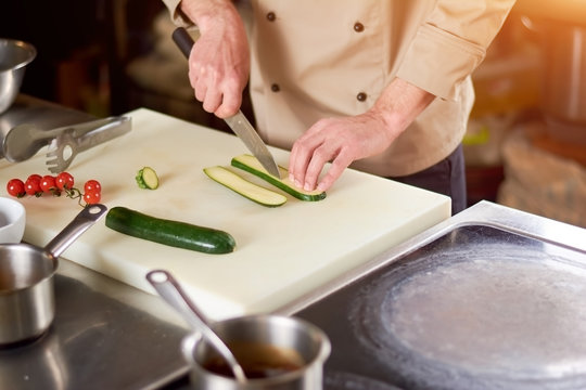 The process of cutting zucchini by chef. The chef in restaurant slicing green marrow on white cutting board. Preparation of dish.