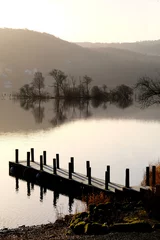  beautiful wooden jetty on lake windemere at sunrise with small island of trees and misty hills in the background © Gill