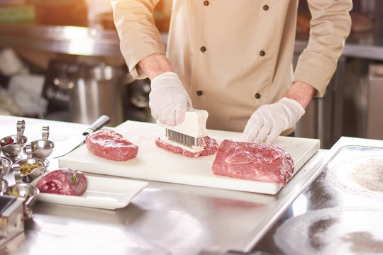 Chef making steak from raw meat. Male chef pressing beef meat on white board at kitchen. Chef cooking steak at professional kitchen.