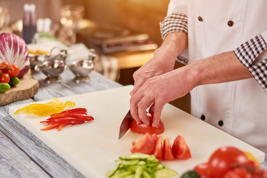 Chef slicing tomato on cutting board. Male chef hands cutting fresh vegetables for salad. Chef at work, kitchen.
