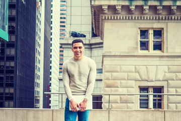 Young American Graduate Student studying in New York, wearing collarless knit sweater, jeans, holding laptop computer, standing outside office, smiling. High buildings on background.