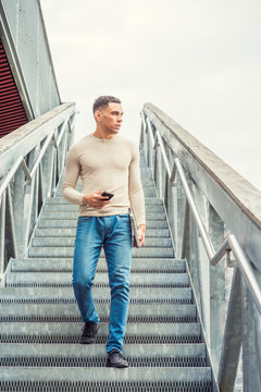 Young American Graduate Student Studying In New York, Wearing Collarless Knit Sweater, Jeans, Sneakers, Carrying Laptop Computer, Holding Cell Phone, Looking Away, Walking Down Stairs On Campus..