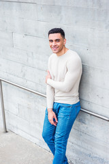Young handsome American Man wearing light gray collarless, knit pullover sweater, blue jeans, standing by wall in New York in spring day, crossing arm, looking up, smiling..