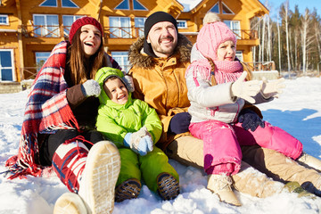 Young Caucasian couple with little kids sitting in snow outdoors, enjoying bright sun and smiling happily