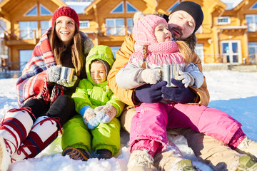 Young Caucasian couple with little children sitting in snow, drinking hot tea and enjoying bright winter sun